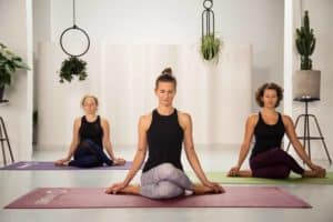 3 junge Frauen sitzen in hellem Raum auf bunten Yogamatten im Schneidersitz und geschlossenen Augen