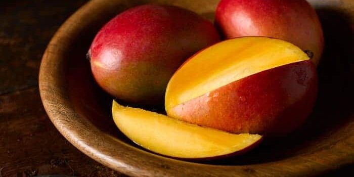 Eine in Scheiben geschnittene Mango, dahinter zwei ganze, rote Mangos