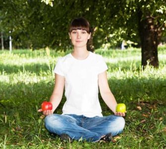 Frau in Jeans und weißem Tshirt sitz im Schneidersitz auf grünem Rasen mit einem roten Apfel in der rechten und einem grünen Apfel in der anderen Hand.