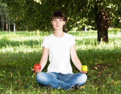 Frau in Jeans und weißem Tshirt sitz im Schneidersitz auf grünem Rasen mit einem roten Apfel in der rechten und einem grünen Apfel in der anderen Hand.