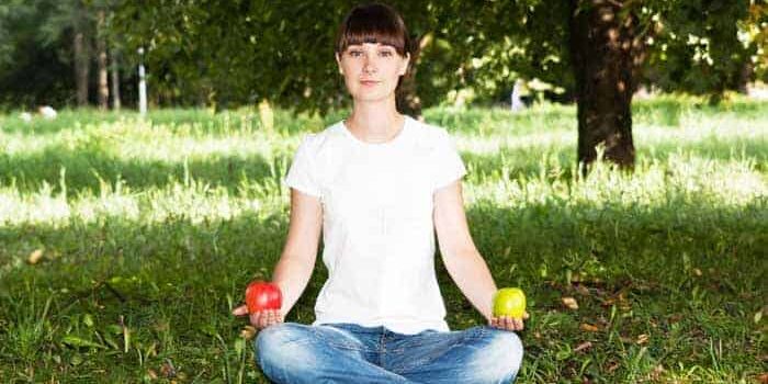 Frau in Jeans und weißem Tshirt sitz im Schneidersitz auf grünem Rasen mit einem roten Apfel in der rechten und einem grünen Apfel in der anderen Hand.
