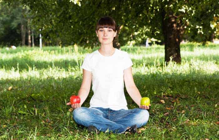 Frau in Jeans und weißem T-Shirt sitz im Schneidersitz auf grünem Rasen mit einem roten Apfel in der rechten und einem grünen Apfel in der anderen Hand.