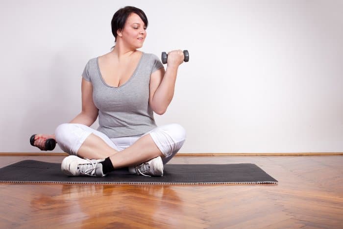 Übergewichtige junge Frau mit dunklen Haaren sitzt in Schneidersitz auf einer Sportmatte und macht einen Bizeps Curl mit Kurzhanteln