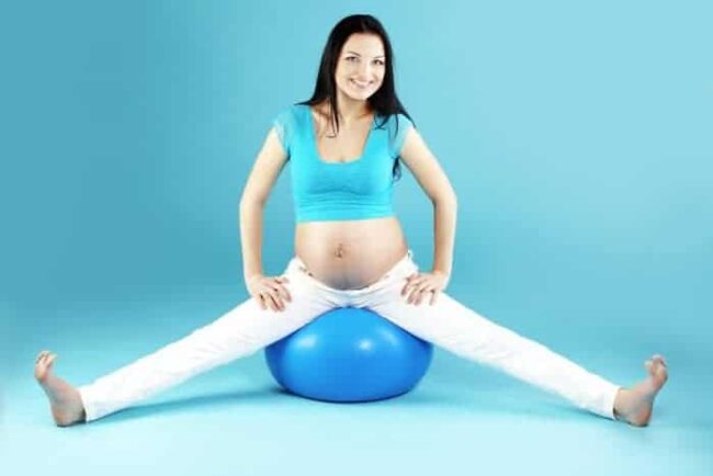 Junge, schwangere Frau mit braunen Haaren und weißer Hode sitzt auf einem blauen Pezziball vor türkisem Hintergrund