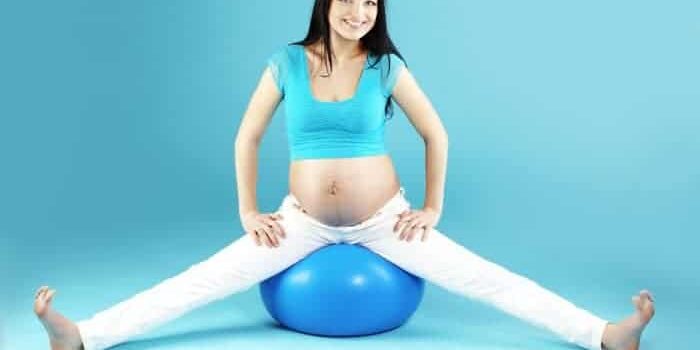 Trainingschwangerschaft Junge, schwangere Frau mit braunen Haaren und weißer Hode sitzt auf einem blauen Pezziball vor türkisem Hintergrund