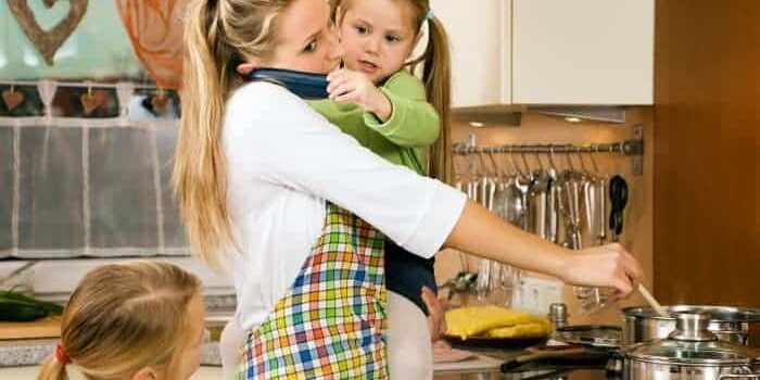 Mutter mit Kind auf dem Arm kocht in der Küche. Zweite Tochter steht neben Ihr. Kind auf dem Arm der Mutter hält der Mutter ein Telefon ans Ohr.
