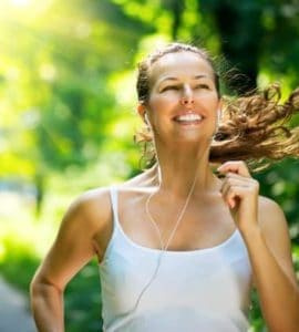 Junge Frau mit weißem Trägertop joggt durch den Wald, lacht und hört Musik.