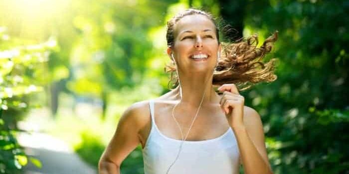 Sporthappy Junge Frau mit weißem Trägertop joggt durch den Wald, lacht und hört Musik.