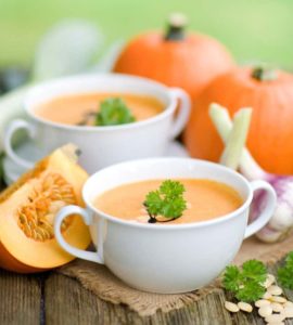 Kürbissuppe in weißer Suppentasse serviert, arrangiert neben Kürbissen und Zwiebeln