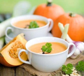 Kürbissuppe in weißer Suppentasse serviert, arrangiert neben Kürbissen und Zwiebeln