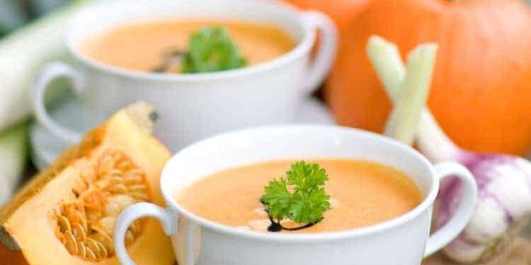 Kürbissuppe in weißer Suppentasse serviert, arrangiert neben Kürbissen und Zwiebeln