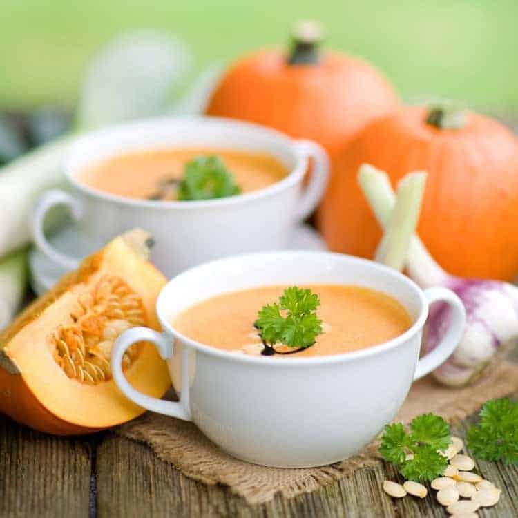 Kürbissuppe in weißer Suppentasse serviert, arrangiert neben Kürbissen und Zwiebeln