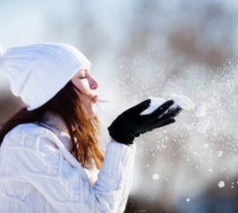 Frau mit weißer Mütze und Schal pustet Schnee aus ihren Händen