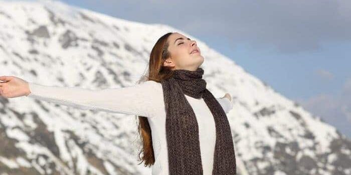 frische-luft-im-winter Frau in weißem Pullover und mit braunem Schal breitet mit geschlossenen Augen die Arme aus und atmet tief ein