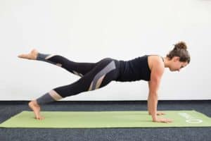 Junge Frau in Liegestützposition auf einer hellgrünen Trainingsmatte macht Pilates Leg Pull Front