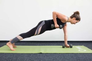 Junge Frau in Liegestützposition auf einer hellgrünen Trainingsmatte zieht mit Kurzhanteln einen Arm hoch