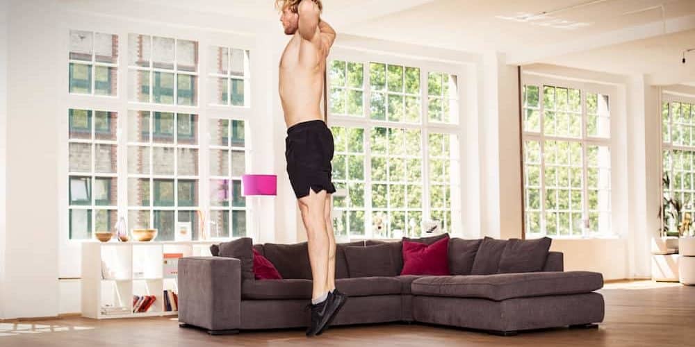 Burpee Strecksprung Mann in heller Wohnung auf dunkler Trainingsmatte macht einen Burpee und einen Strecksprung mit Händen hinter dem Kopf