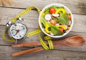 Eine Uhr, ein Maßband und eine Schale Salat verdeutlichen, dass man bei Intervall Fasten nur zu bestimmten Uhrzeiten isst, um Abzunehmen