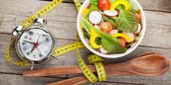 Eine Uhr, ein Maßband und eine Schale Salat verdeutlichen, dass man bei Intervall Fasten nur zu bestimmten Uhrzeiten isst, um Abzunehmen