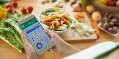 Frau hat Smartphone in den Händen mit App zum Kalorienzählen, in Hintergrund bunte Lebensmittel auf Holztisch
