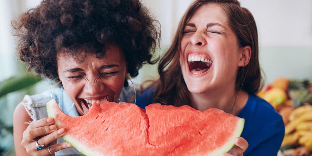 Teller2 Zwei junge Frauen lachen und essen zusammen ein großes Stück Wassermelone