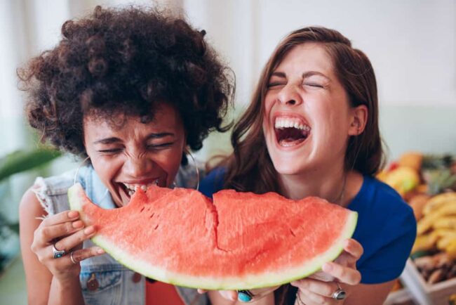 Teller2 Zwei junge Frauen lachen und essen zusammen ein großes Stück Wassermelone