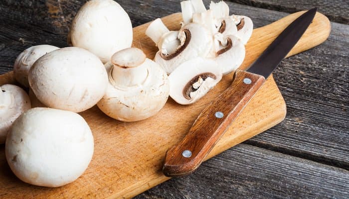 Weiße Champignons auf Holzbrett neben einem Messer mit Holzgriff