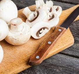 Weiße Champignons und ein Messer auf einem Holzbrett, zum Teil am Stück, zum Teil geschnitten