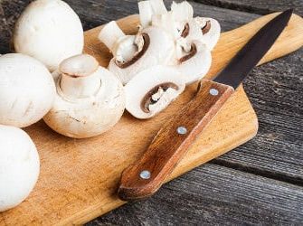 Weiße Champignons und ein Messer auf einem Holzbrett, zum Teil am Stück, zum Teil geschnitten