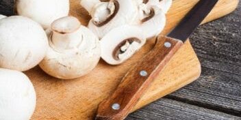 Weiße Champignons und ein Messer auf einem Holzbrett, zum Teil am Stück, zum Teil geschnitten