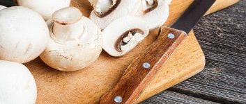 Weiße Champignons und ein Messer auf einem Holzbrett, zum Teil am Stück, zum Teil geschnitten