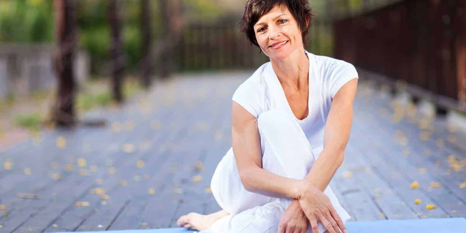Problemzonen ab 50 Frau Mitte 50 in weißer Sportkleidung sitzt auf blauer Trainingsmatte und lächelt glücklich und entspannt