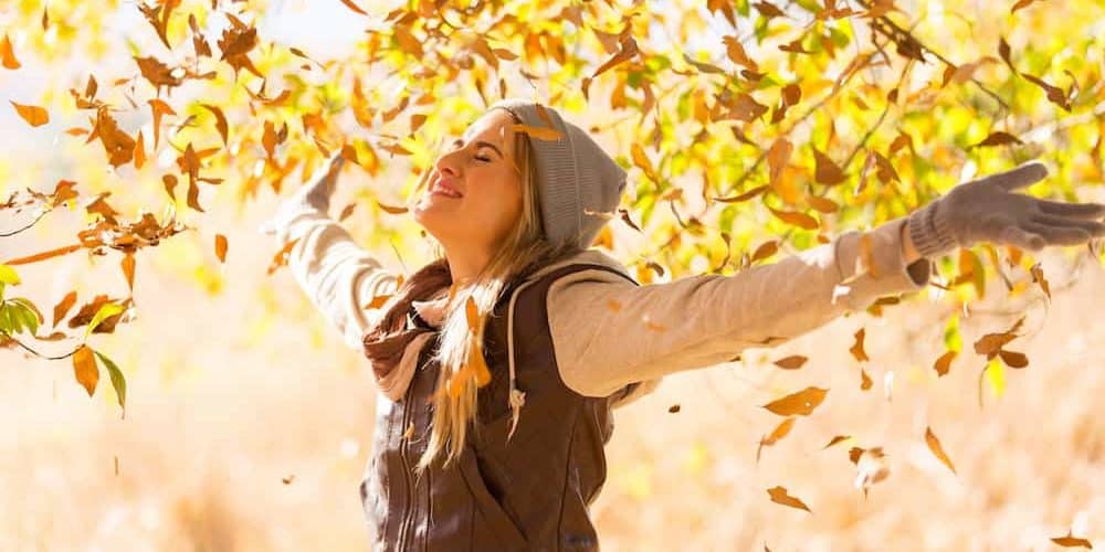Gute Laune im Herbst Junge Frau mit Jacke und Mütze streckt die Arme nach oben und ist glücklich. Herbst Laub fliegt im Hintergrund durch die Luft