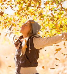 Junge Frau mit Jacke und Mütze streckt die Arme nach oben und ist glücklich. Herbst Laub fliegt im Hintergrund durch die Luft