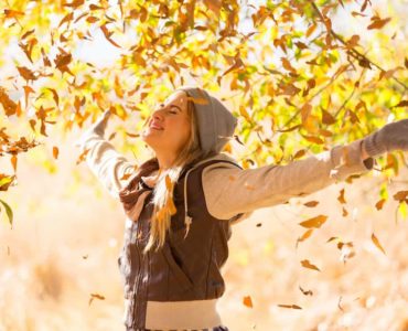 Junge Frau mit Jacke und Mütze streckt die Arme nach oben und ist glücklich. Herbst Laub fliegt im Hintergrund durch die Luft