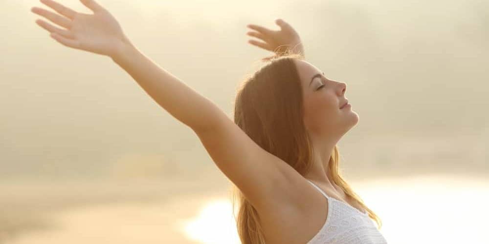 Junge Frau am Meer atmet tief durch und breitet dabei die Arme aus