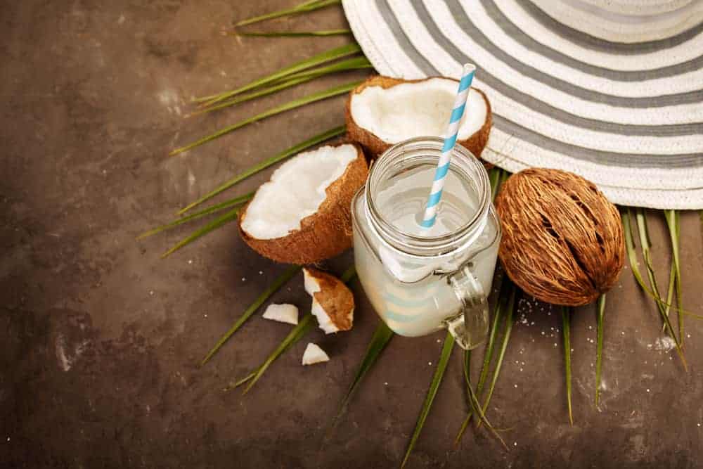 Glas mit Kokoswasser und Strohhalm zwischen frischen Kokosnüssen auf dunklem Holztisch