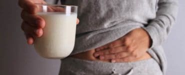 Frau mit Milchglas in der Hand fasst sich an den Bauch, hat Blähungen durch Laktoseintoleranz