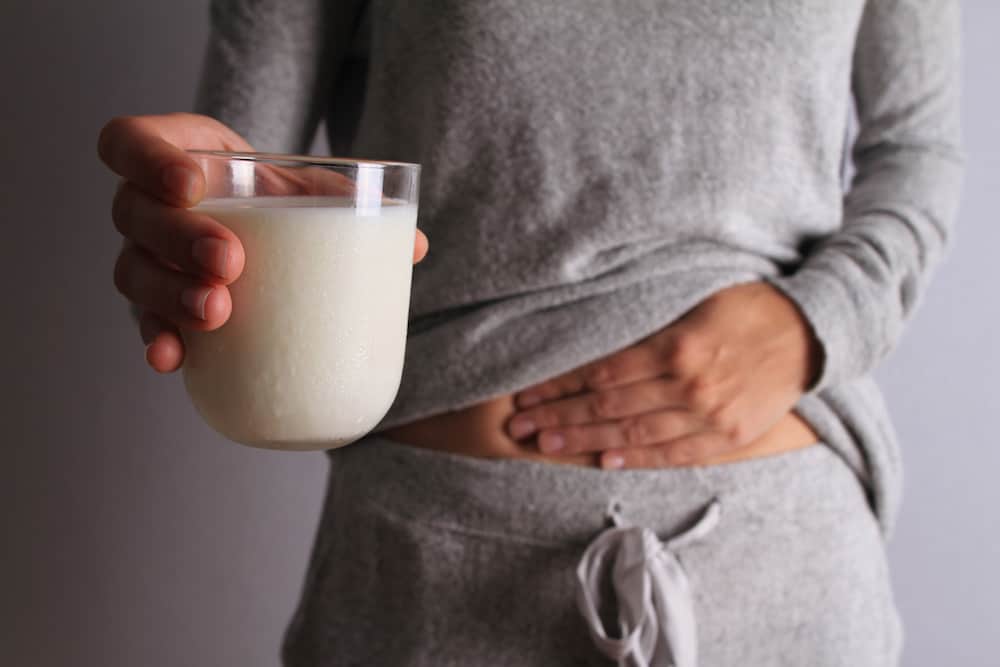 Frau mit Milchglas in der Hand fasst sich an den Bauch, hat Blähungen durch Laktoseintoleranz