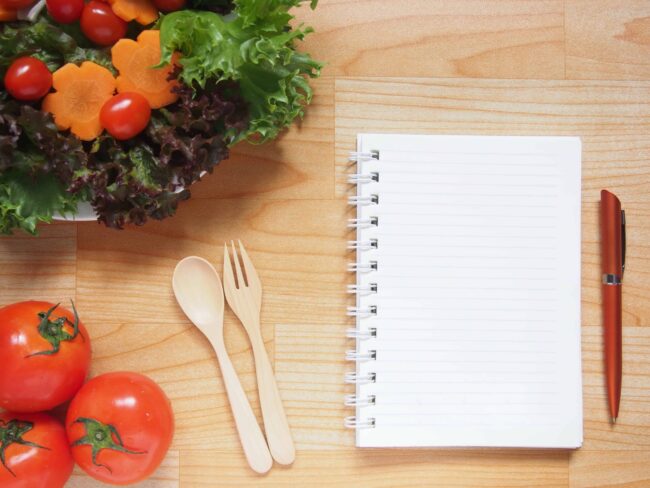 Holztisch mit gesundem frischem Gemüse und einem unbeschriebenen Ringblock mit Kuli daneben