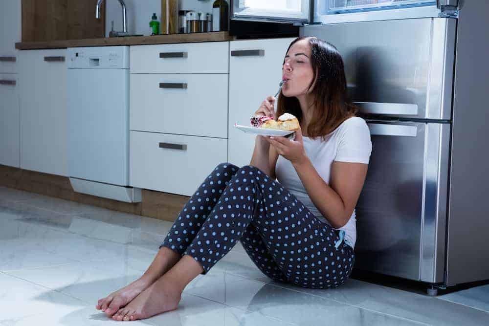 Junge Frau im Pyjama sitzt nachts in der Küche an den Kühlschrank gelehnt und ist Torte