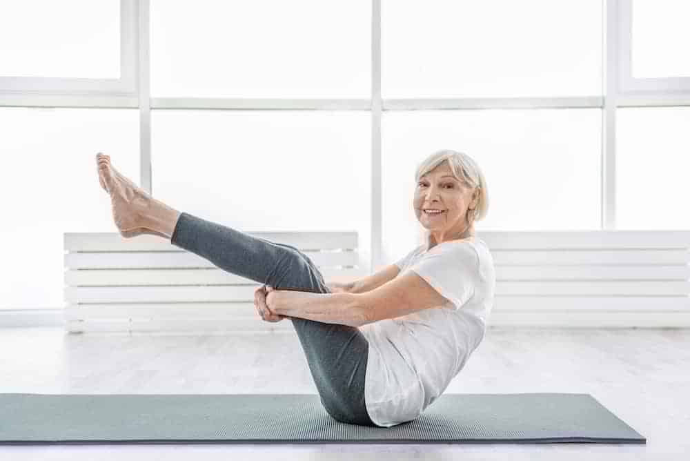 Ältere fitte Frau in Sportkleidung macht Yoga Pose Boot in hellem Raum auf grauer Matte