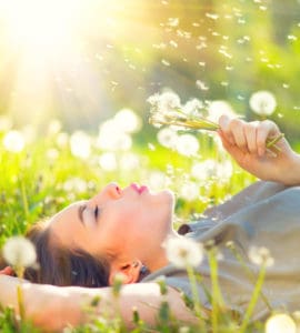 Junge Frau genießt den Moment liegt auf einer Wiese und pustet gegen eine Pusteblume.