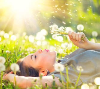 Junge Frau genießt den Moment liegt auf einer Wiese und pustet gegen eine Pusteblume.