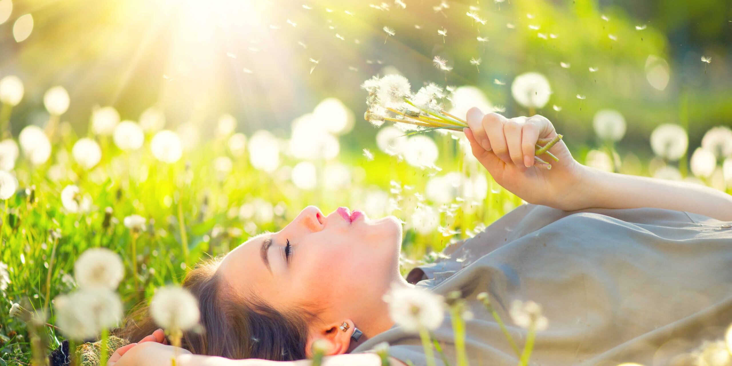 Junge Frau genießt den Moment liegt auf einer Wiese und pustet gegen eine Pusteblume.
