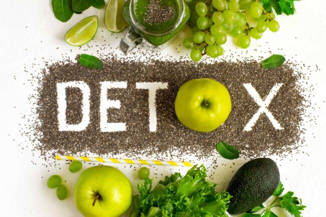 Tischplatte in der Mitte steht das Wort DETOX, darüber und darunter grünes Gemüse und Obst
