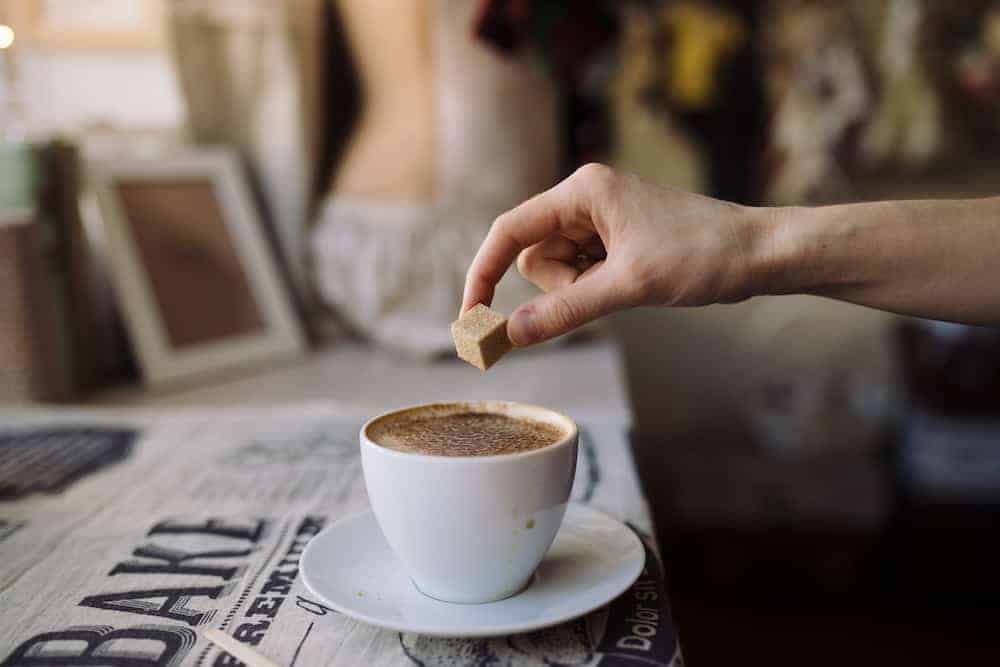Hand einer jungen Frau wirft einen Würfelzucker in eine Kaffeetasse mit Capuccino