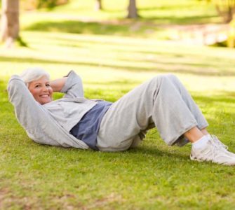 Ältere fitte Frau in Sportkleidung im Park liegt auf Wiese und macht Crunches
