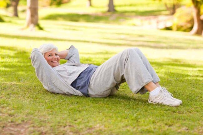 Ältere fitte Frau in Sportkleidung im Park liegt auf Wiese und macht Crunches